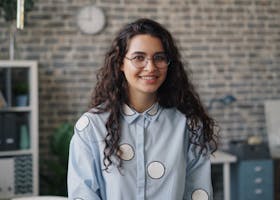 Portrait of a smiling woman with curly hair and eyeglasses in an office setting.